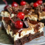 Close-up of a brownie cheesecake square topped with whipped cream, chocolate shavings, and a cherry.