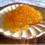 A small white plate holds a mound of bright yellow passion fruit caviar on a red and white checkered tablecloth.