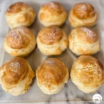 Six golden-brown cream puffs arranged on a white wooden surface.
