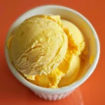 A scoop of creamy orange sherbet in a glass bowl sits on a red background.